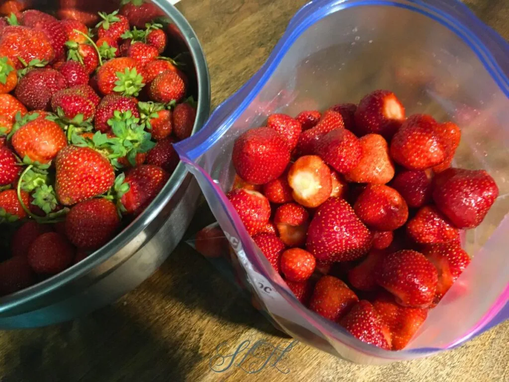 metal mixing bowl of fresh strawberries next to a ziplock plastic bag of clean whole strawberries with the stems taken of on a wooden counter
