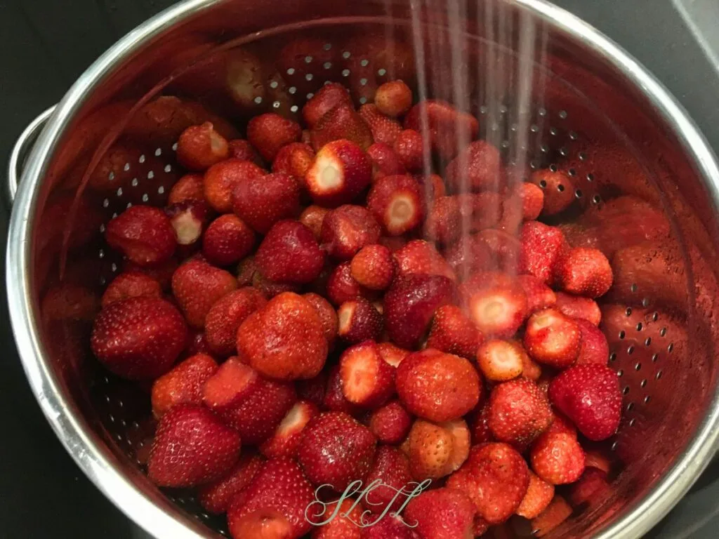 fresh strawberries with stems taken off being rinsed under water in a colander 