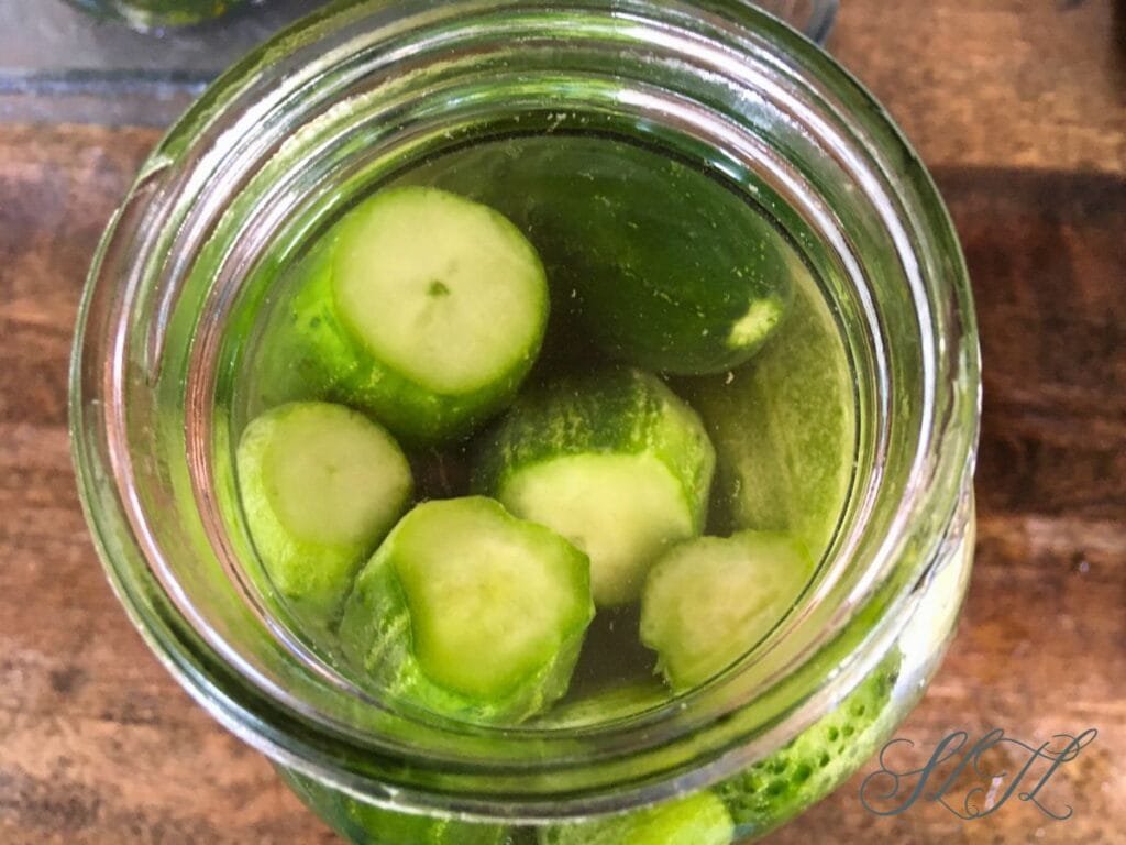 canning jar packed with raw cucumbers and fermenting salt brine with 2 cucumbers sitting on a wooden counter