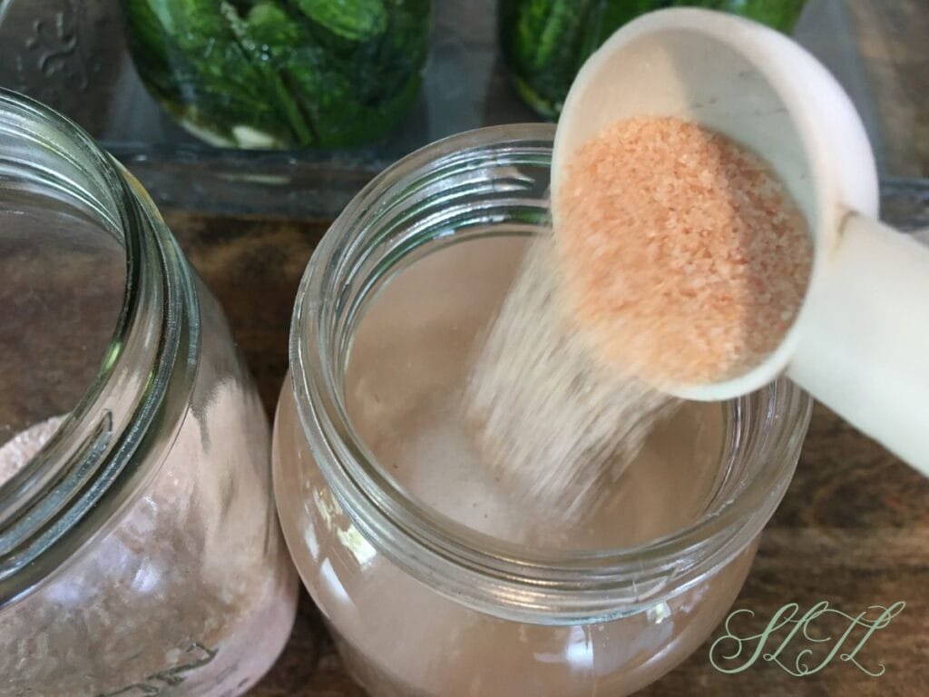 Himalayan salt being poured into a quart jar of water, a jar of salt sitting next to it with fermenting pickles in the background.