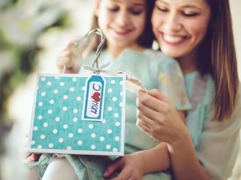 girl sitting on mom's lap smiling as mom looks at a note on a blue and white polkadot gift bag