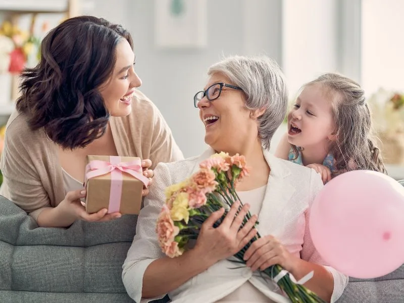 grandma sitting on a couch holding flowers being given a gift box by her daughter and a pink balloon by her granddaughter.