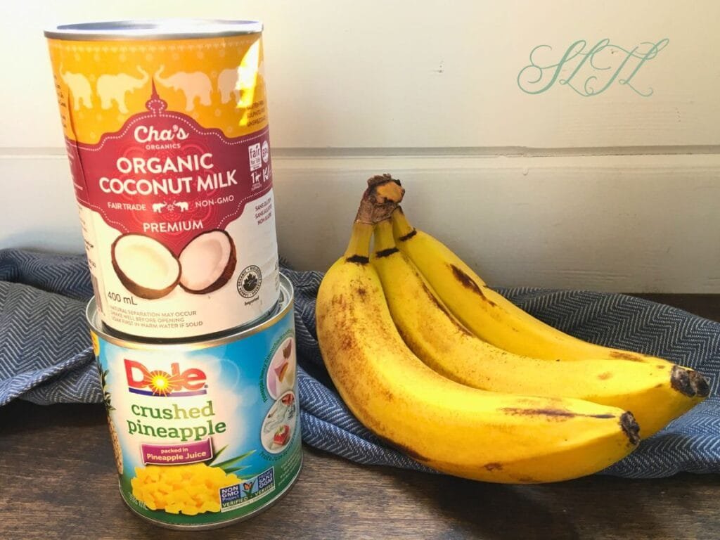 bananas, canned coconut milk and canned pineapple on wooden counter with blue tea towel.