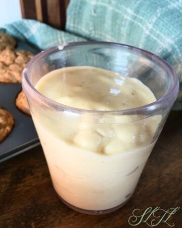 Pinna colada smoothie in clear cup with muffins, wooden cutting board and tea towel in background.