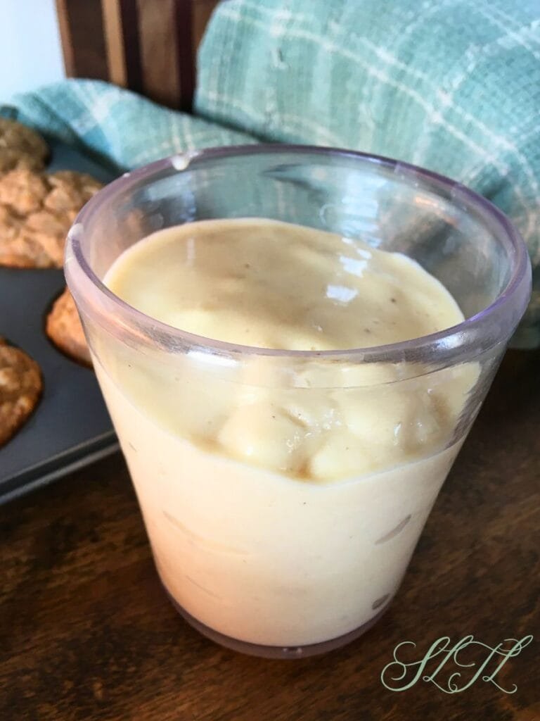 Pinna colada smoothie in clear cup with muffins, wooden cutting board and tea towel in background.
