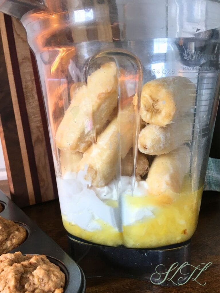 Frozen bananas, coconut and pineapple in blender next to a pan of muffins, and in front of a wooden cutting board and teal tea towel.