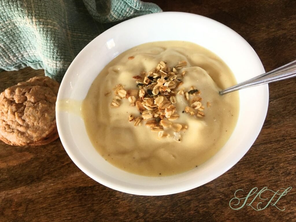 Pinna colada smoothie bowl topped with granola with spoon in the bowl. Sitting on wooden table next to a muffin and tea towel.