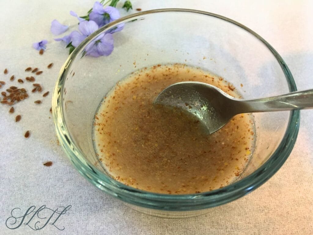 ground flaxseed being mixed into water in glass bowl with flax seeds and flaxseed flower sitting behind it
