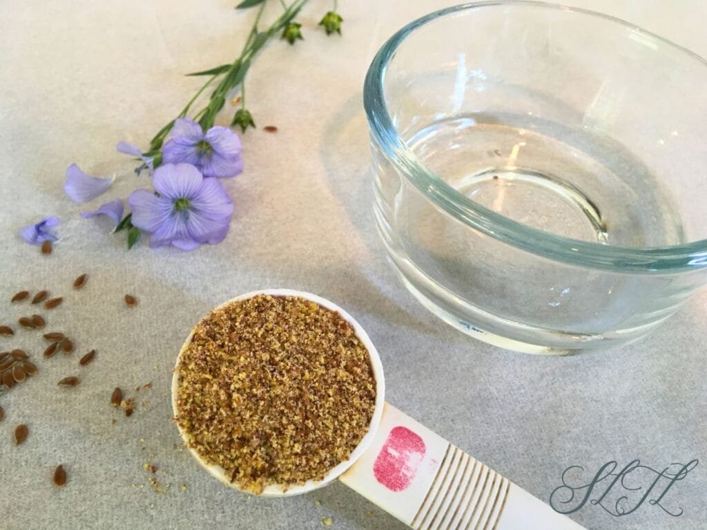 glass bowl of water, one tablespoon full of ground flaxseed, a few flax seeds sprinkled next to it along with flaxseed flower laying beside bowl.