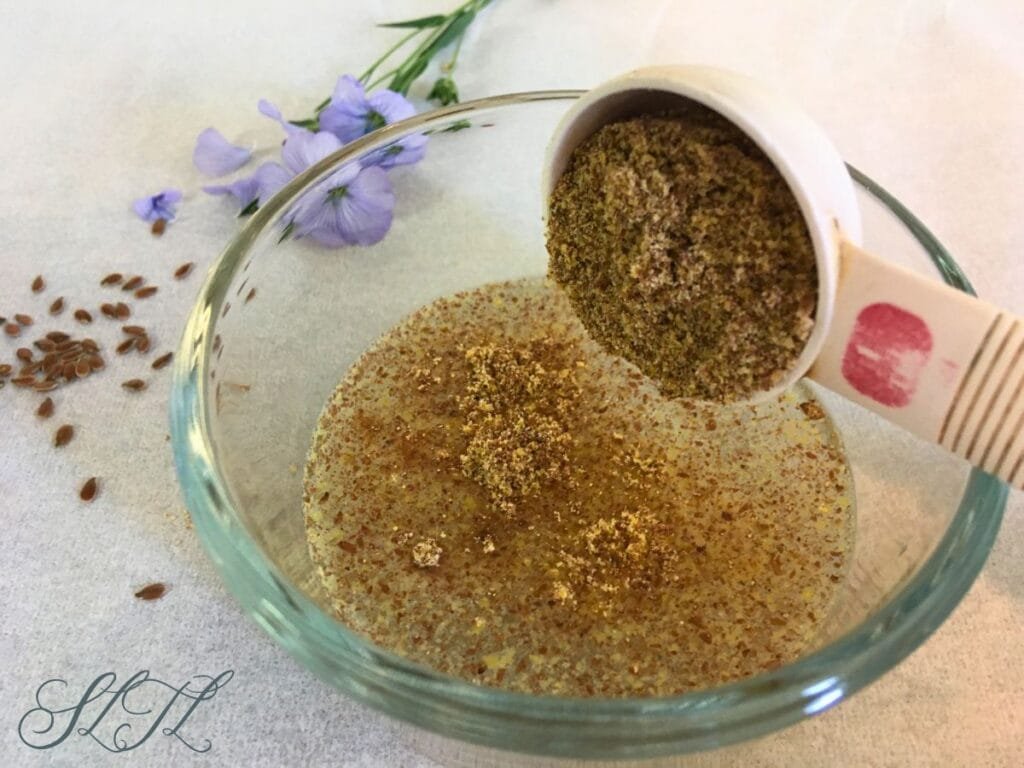 ground flaxseed being poured into water in glass bowl with flax seeds and flaxseed flower sitting in back ground