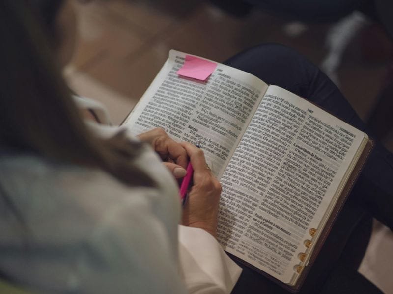 over the shoulder of woman reading open Bible on her lap with pink sticky note on top