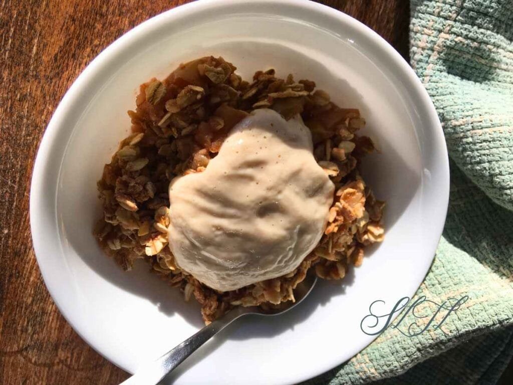 vegan apple rhubarb crisp topped with vegan pear cream in a white bowl with a spoon sitting on a wooden table next to an aqua tea towel