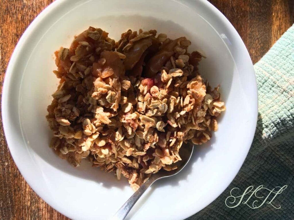 vegan apple rhubarb crisp in a white bowl with a spoon sitting on a wooden table next to an aqua tea towel