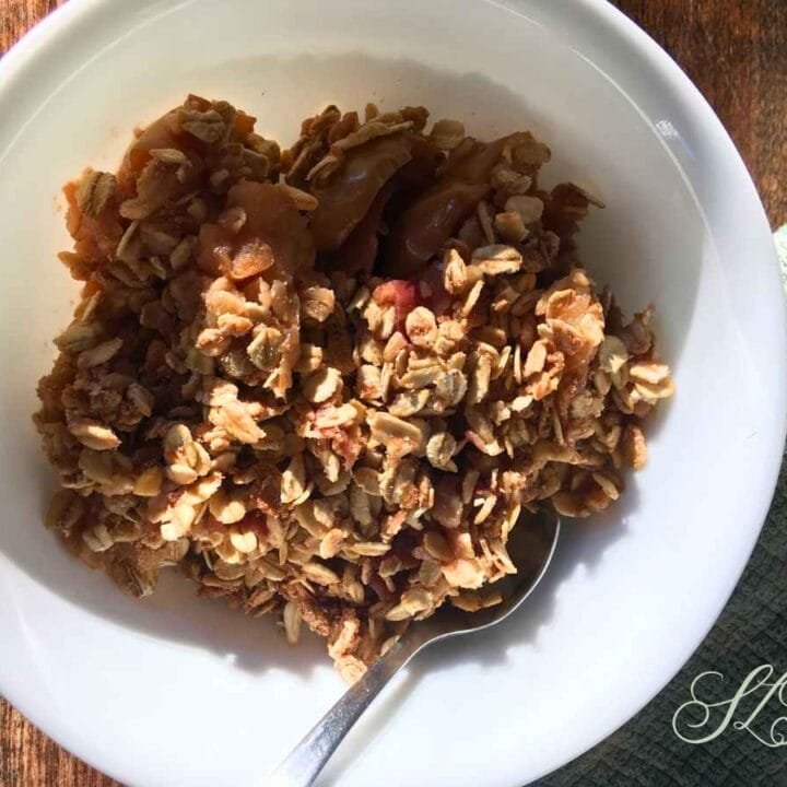 apple crisp with rhubarb in a white bowl with a silver spoon on a wooden table next to a teal tea towel