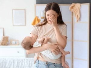 Tired, stressed and overwhelmed young mom with hand on her head holding a sleeping baby in the other arm.