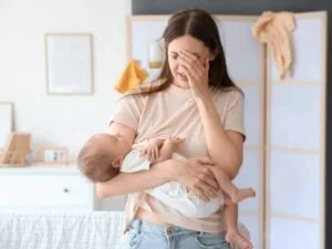 Tired, stressed and overwhelmed young mom with hand on her head holding a sleeping baby in the other arm.