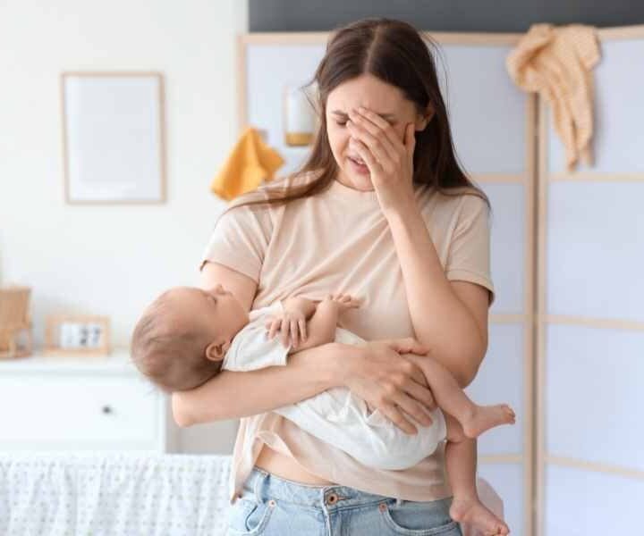 Tired, stressed and overwhelmed young mom with hand on her head holding a sleeping baby in the other arm.