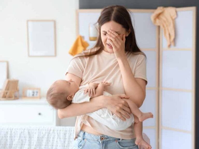 Tired, stressed and overwhelmed young mom with hand on her head holding a sleeping baby in the other arm.